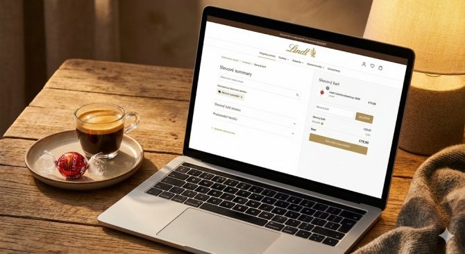 Detailný záber na obrazovku notebooku s otvoreným košíkom e-shopu Lindt a poľom pre zadanie zľavového kódu, vedľa šálky kávy a rozbalenej pralinky Lindor.
